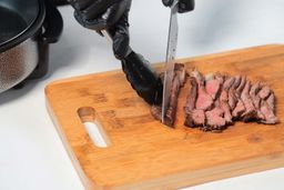 On the cutting board, a tong is holding the steak while a knife is slicing the steak.