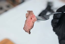 A hand in black glove using a fork to pick up a slice of steak. In the background is the Ovente Non-Stick Electric Skillet SK11112B.