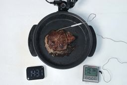 A piece of deep golden brown steak inside the Ovente Non-Stick Electric Skillet SK11112B. In front of the skillet is a digital timer and a meat thermometer with its probe inserted into the steak.