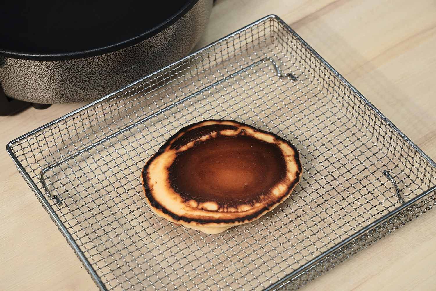 The top side of a dark brown pancake with highly noticeable dark edges inside an air fryer basket. In the corner is the Ovente Non-Stick Electric Skillet SK11112B.