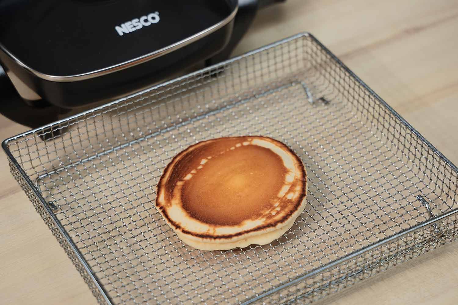 The top side of a mottled golden brown pancake with noticeable dark edges inside an air fryer basket. In the corner is the Nesco Non-Stick Electric Skillet ES-08.