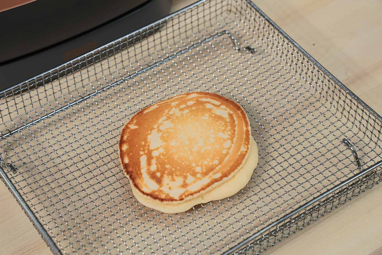 The top side of a mottled golden brown pancake that’s dark around the edges inside an air fryer basket. In the corner is the Crux Non-Stick Electric Skillet 14620.