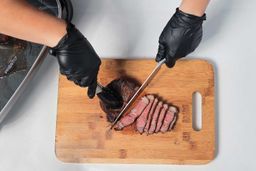 On the cutting board, a tong is holding the steak while a knife is slicing the steak.