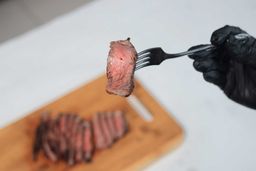 A hand in black glove using a fork to pick up a slice of steak.