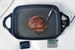 A piece of golden brown steak inside the Presto Electric Skillet 06852. In front of the skillet is a digital timer and a meat thermometer with its probe inserted into the steak.