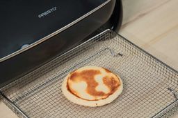 The bottom side of a golden brown pancake inside an air fryer basket. In the corner is the Presto Electric Skillet 06852.