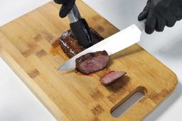 On the cutting board, a tong is holding the steak while a knife is slicing the steak.