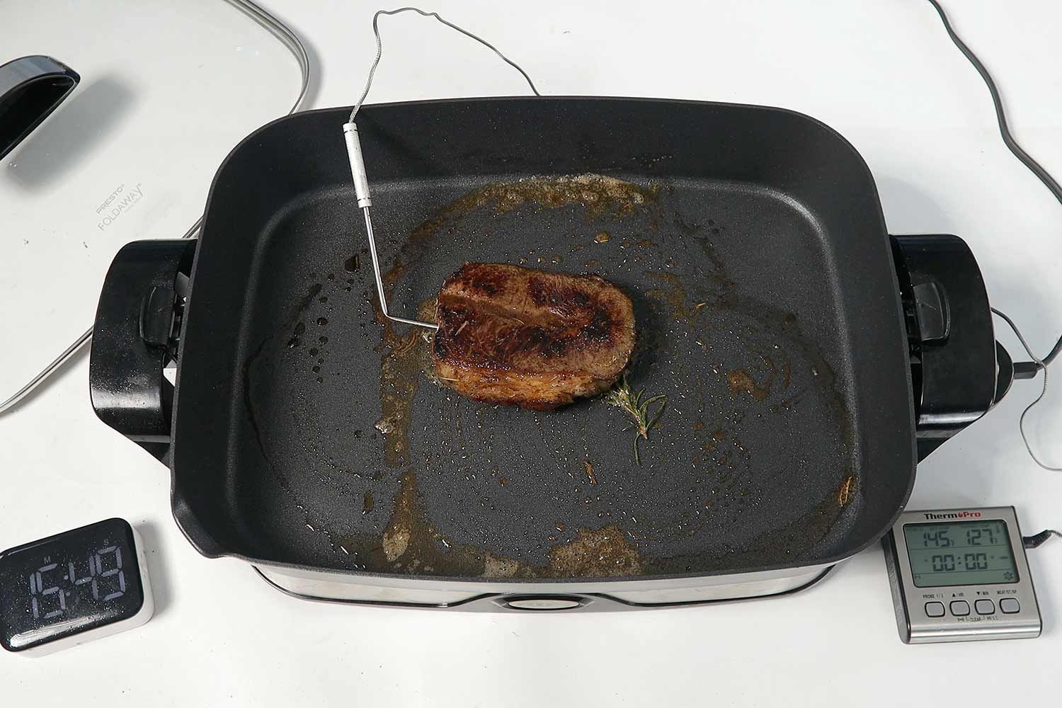 A piece of golden brown steak inside the Presto Foldaway Non-Stick Electric Skillet 06857. In front of the skillet is a digital timer and a meat thermometer with its probe inserted into the steak.