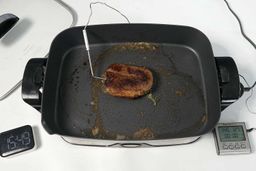 A piece of golden brown steak inside the Presto Foldaway Non-Stick Electric Skillet 06857. In front of the skillet is a digital timer and a meat thermometer with its probe inserted into the steak.