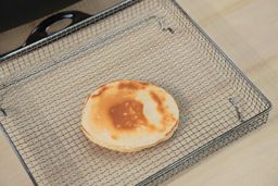 The bottom side of a golden brown pancake inside an air fryer basket. In the corner is the Presto Foldaway Non-Stick Electric Skillet 06857.