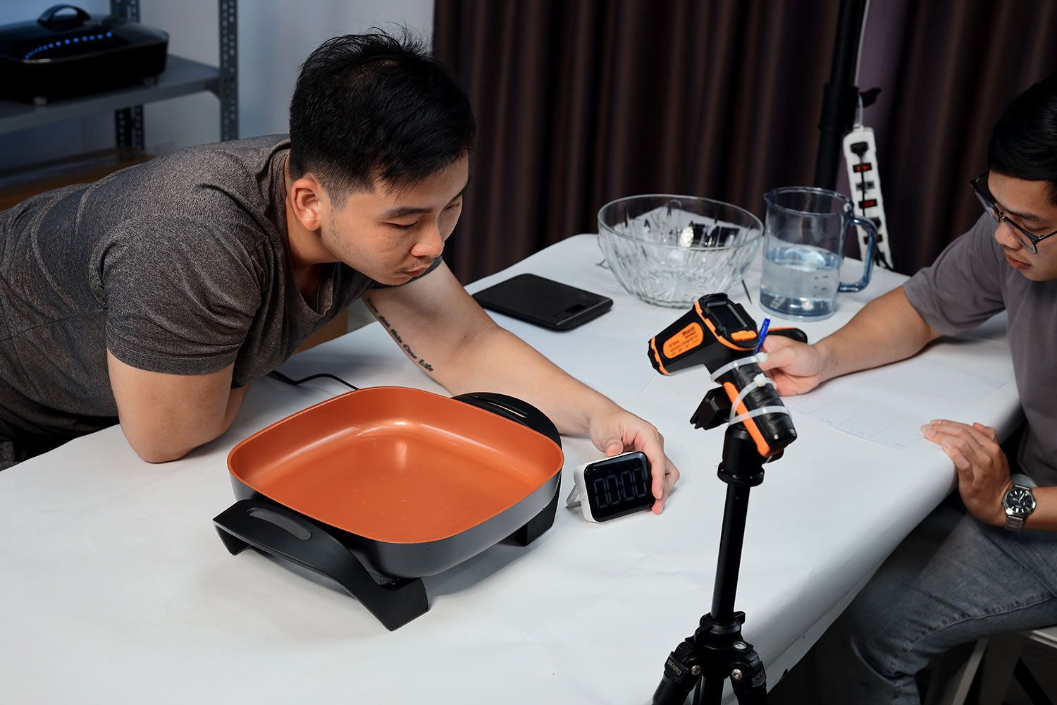 An infrared thermometer fixed on a stand, the Bella Non-Stick Electric Skillet 14607, a person adjusting a digital thermometer, another person holding a pen.