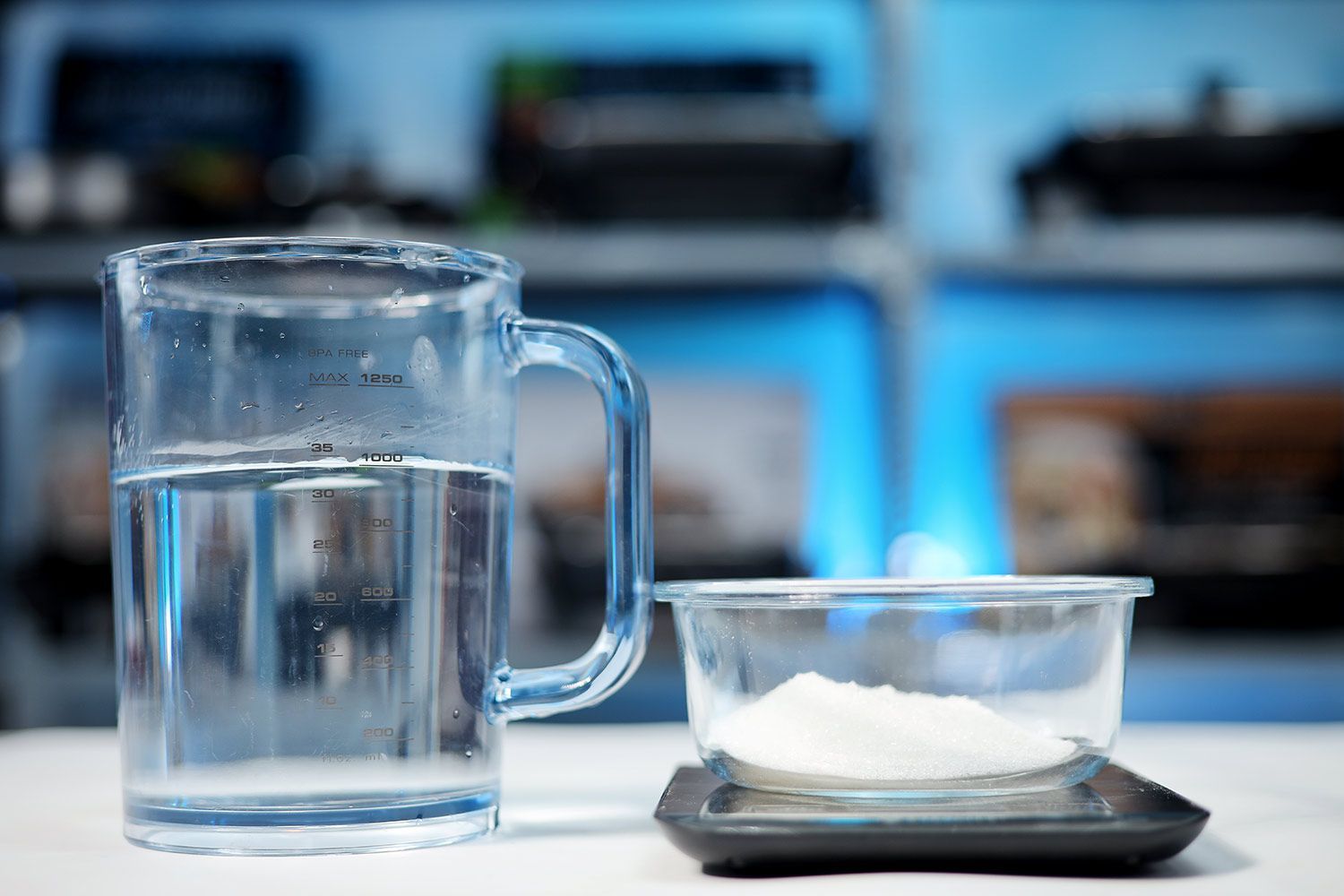 A measuring cup with 1 liter of water and a bowl of sugar on a digital scale.