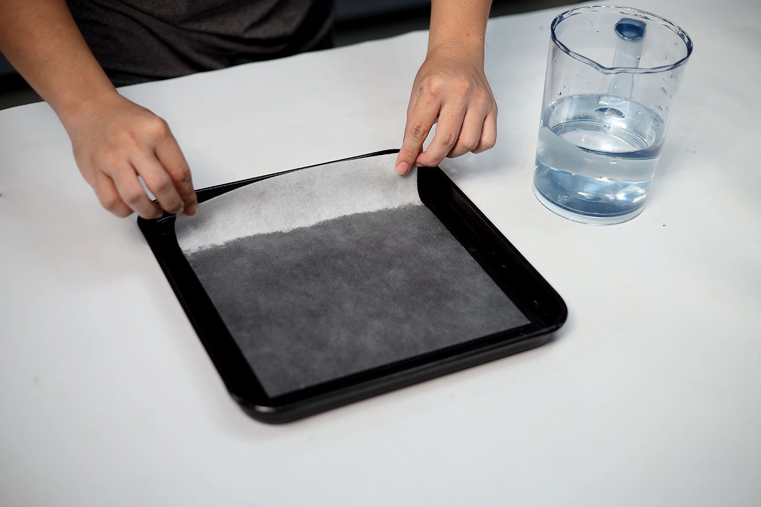 Two hands soaking an oil absorbent sheet into a tray of sugar water. Next to the tray is a measuring cup of sugar water.