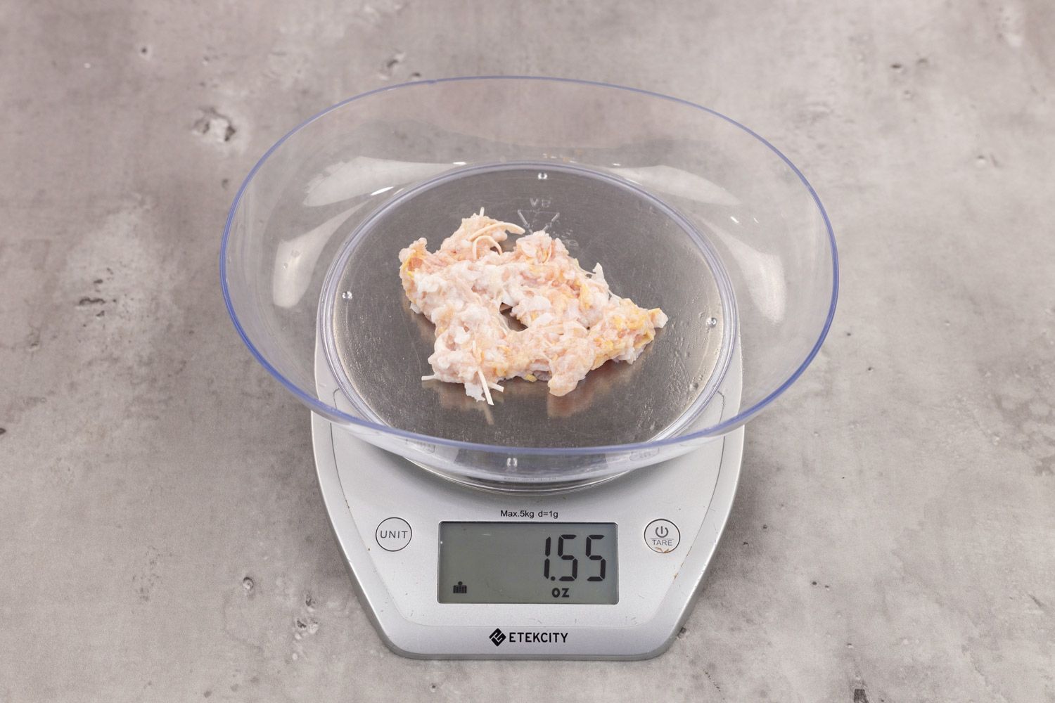 1.55 ounces of ground chicken scraps from garbage disposal, displayed on digital scale, placed on granite-looking table. Visible pieces of shredded tendon and shredded bone among mess of soft tissue.