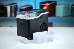 A countertop ice maker with a scoop filled with ice in front and an empty ice basket to the right side.