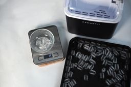 To the right of a portable ice maker is a small bowl with broken ice weighing 19 g. To the right is a baking tray of unbroken ice.