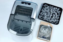 Top down view of the Frigidaire EFIC189 ice maker to the left with one load of ice removed and placed into a black colored baking tray on the right. Badly formed ice is seen separated into a bowl on a scale showing 15 grams.