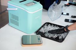 A full basket of ice from a countertop ice maker tipped for sorting into a black making tray with the ice machine in the background and a small scale in the foreground.