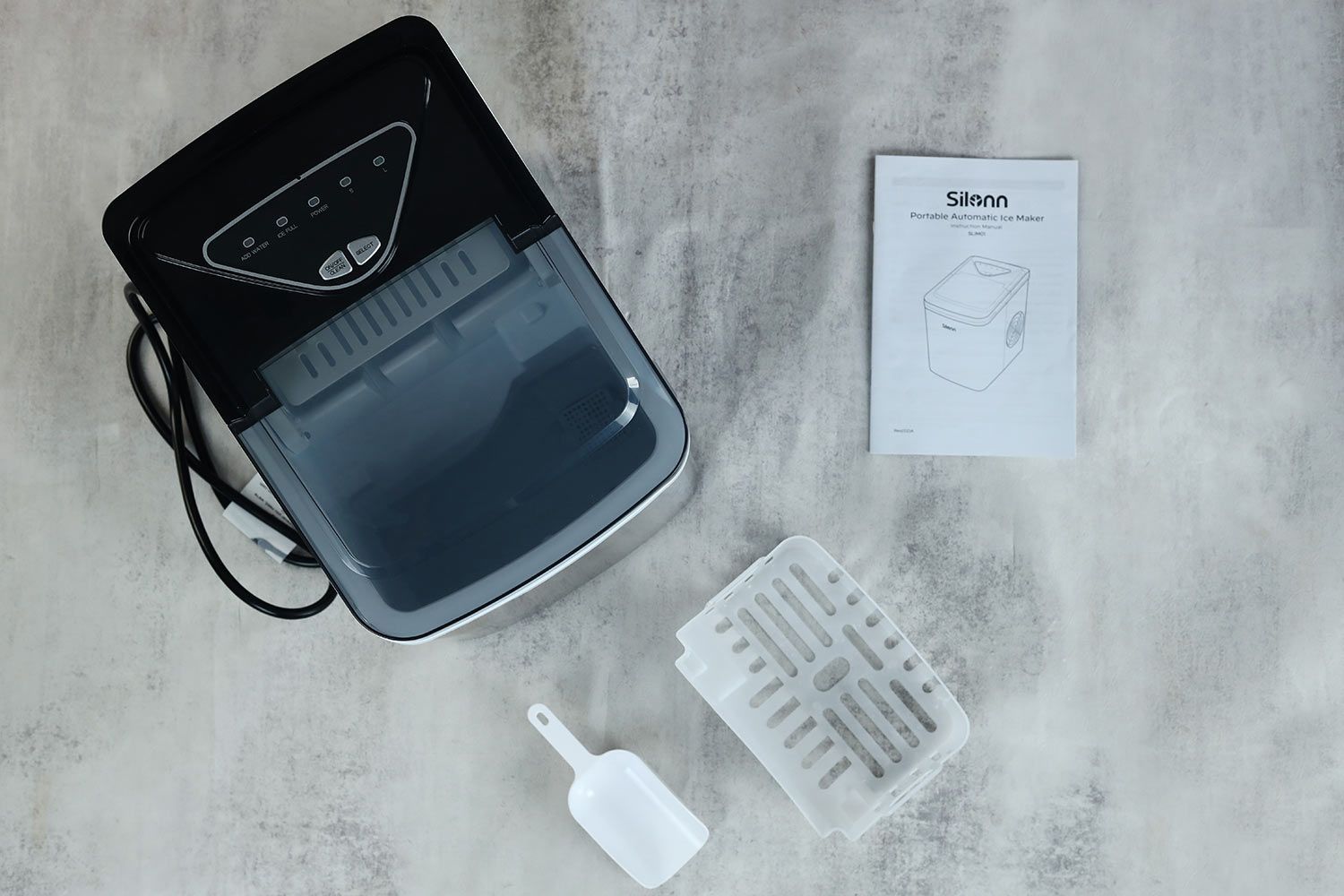 The unboxed Silonn portable ice maker on a gray background with the ice basker and plastic scoop in front of the machine and the user manual set to the rear right.