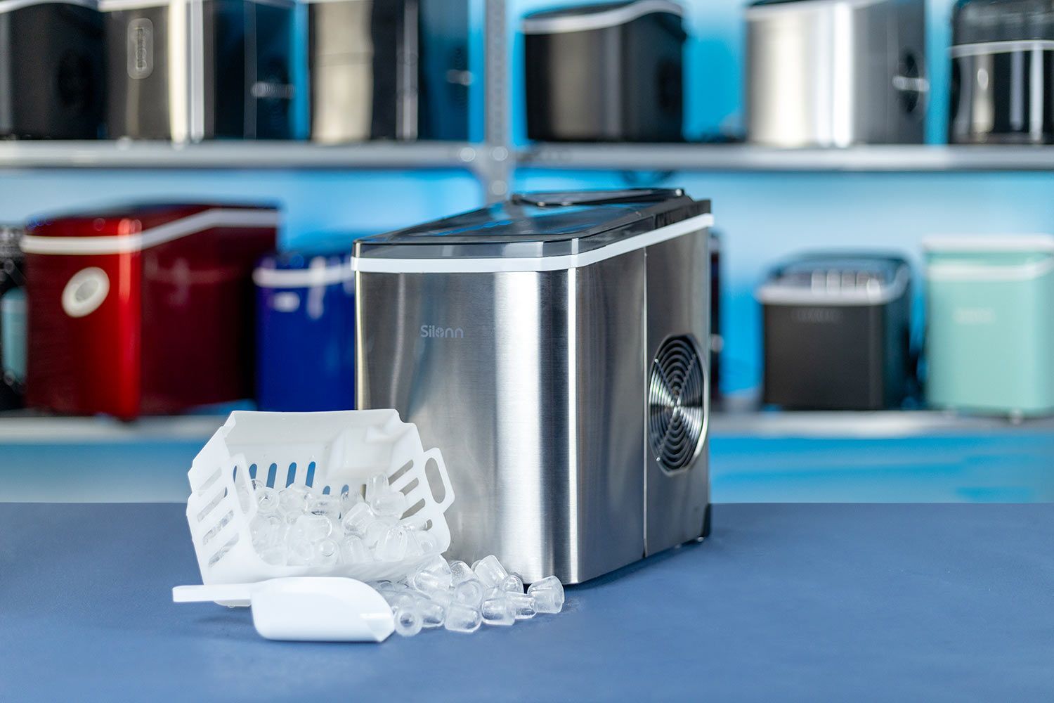 The Silonn Countertop Ice Maker SLIM01 with a basket of ice toppled over in the front and a shelf in the background with numerous other ice makers.