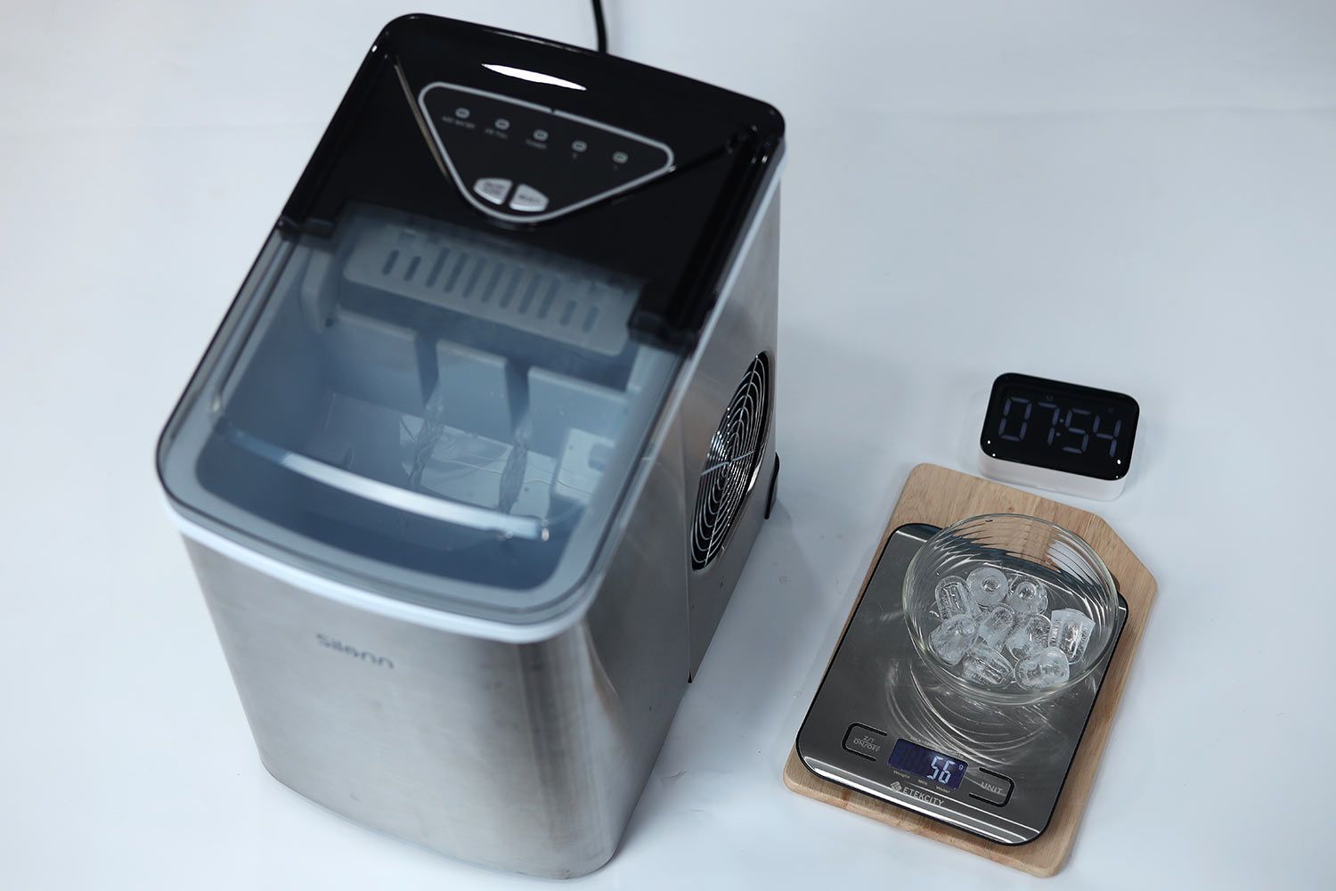 Top down view of an ice maker to the left and on the right on a small wooden board is a scale with a bowl containing 56 grams of ice from one cycle of large ice bullet making.