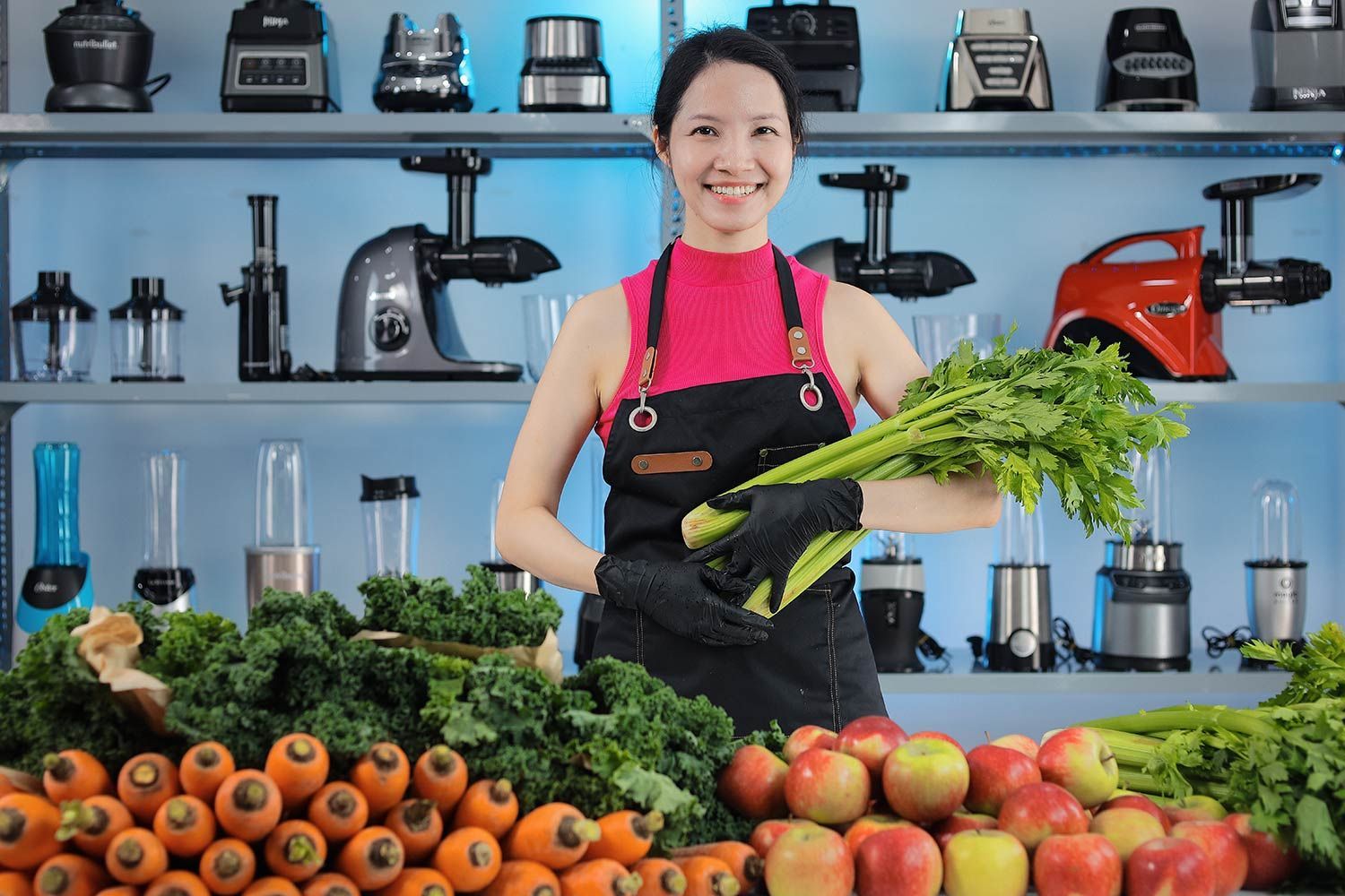 Woman holding celery, with piles of carrots, apples, kale, and celery in front of her