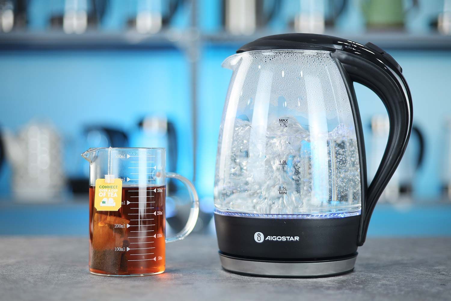 The Aigostar Electric Kettle 300104LCB on the right and a cup of black tea on the left. In the background is a shelf with different electric kettles.