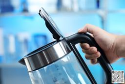 A hand holding the Amazon Basics Electric Glass and Steel Kettle (F-625C) by its handle with its lid open.