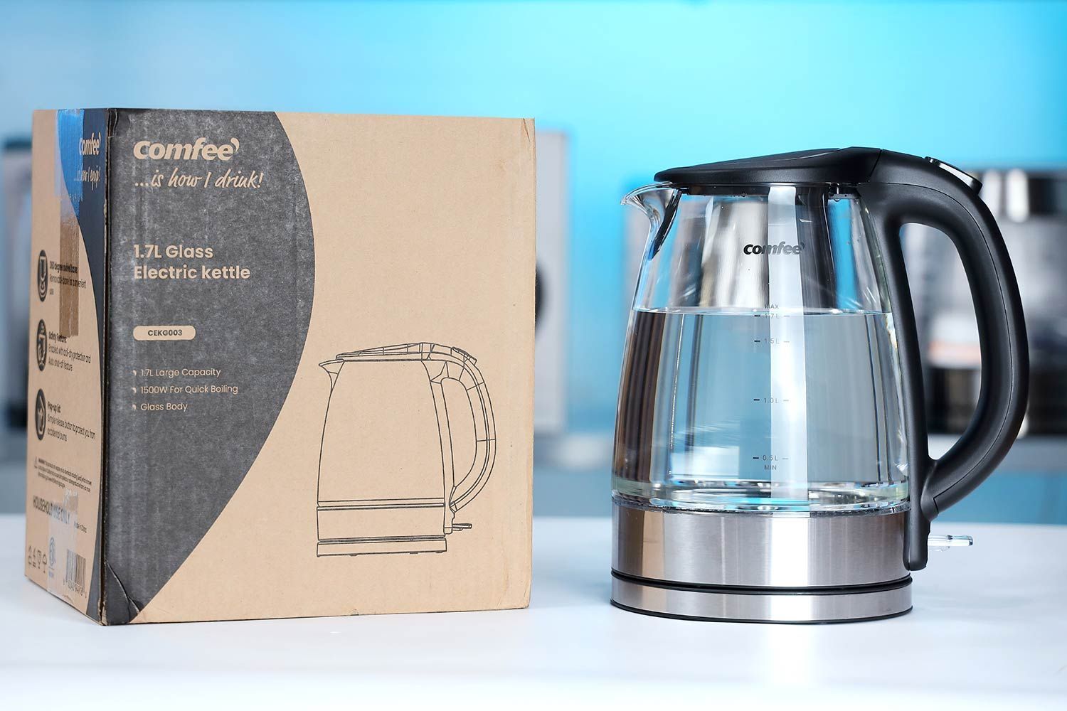 The Comfee Glass Electric Kettle (CEKG003) on the right and its cardboard box on the left. In the background is a shelf with different electric kettles.