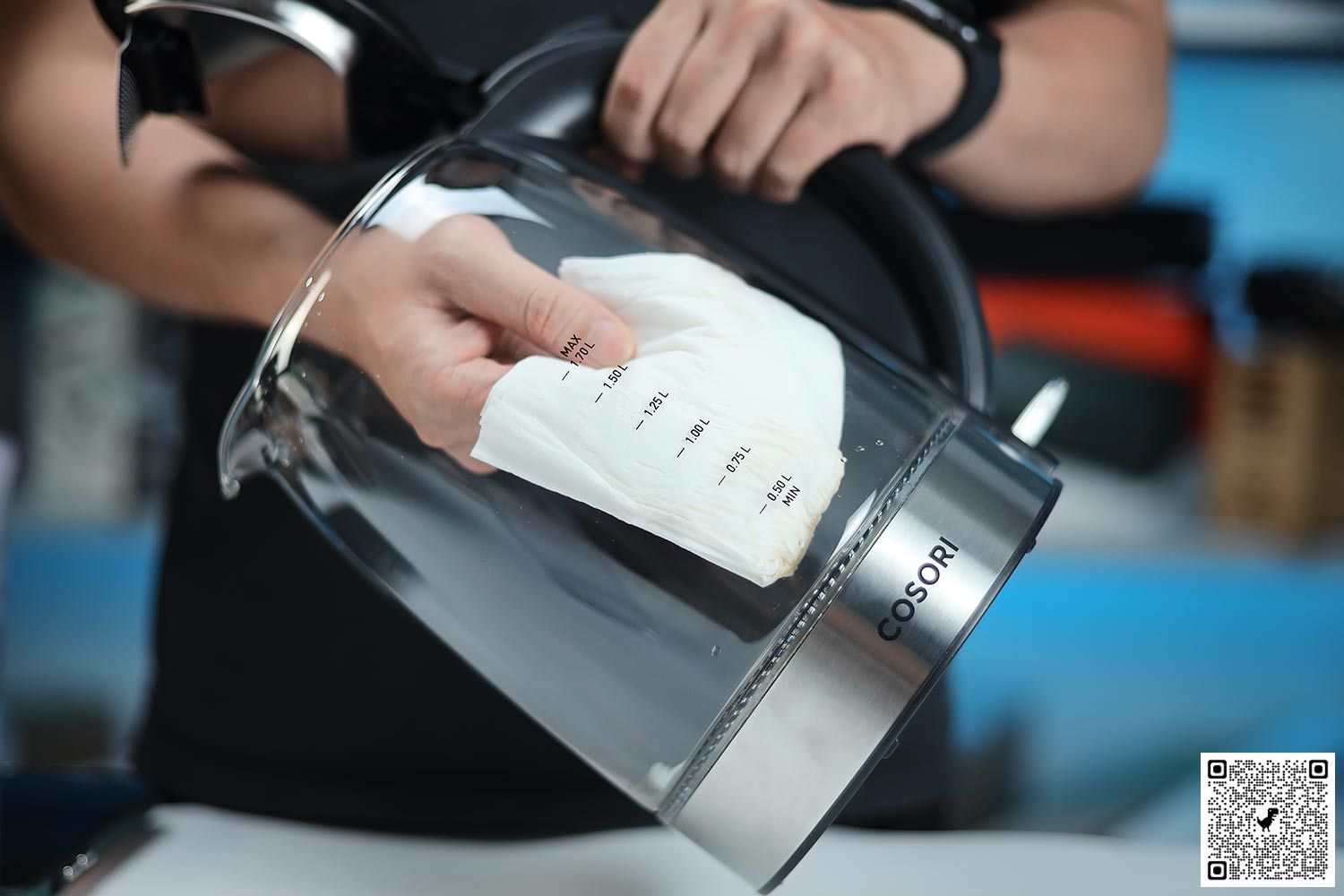 A person in a black shirt holding the Cosori Glass Electric Kettle (GK172-CO) by its handle on one hand and the other hand wiping the carafe interior with a piece of tissue.