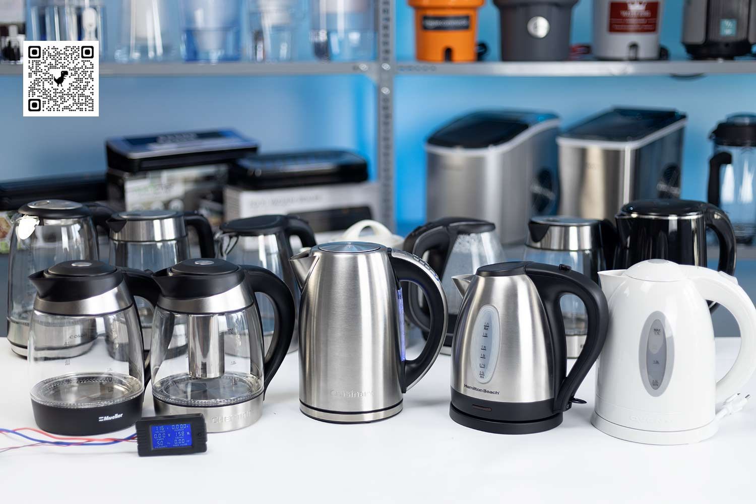 On a white table is 12 electric kettles. In front of them is a grey and blue-screened power meter. 