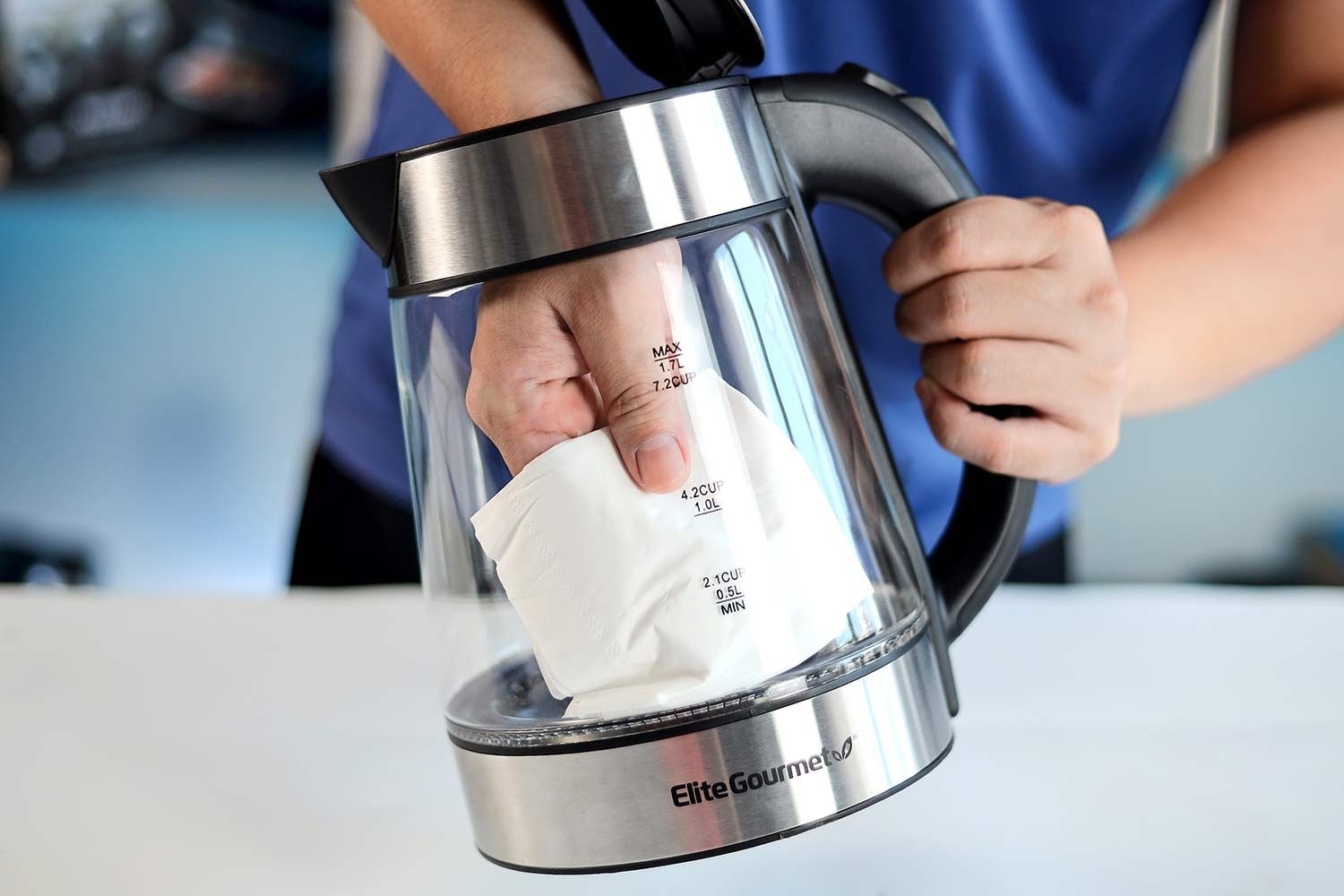 A person in a blue shirt holding the Elite Gourmet Electric Glass Kettle (EKT-602) by its handle on one hand and the other hand wiping the carafe interior with a piece of tissue.