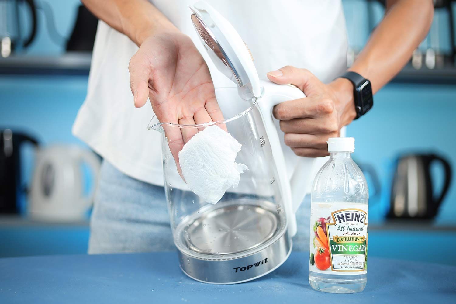 A person in a white shirt holding the Topwit Glass Electric Tea Kettle T630 by its handle on one hand and the other hand wiping the carafe interior with a piece of tissue. On the right is a bottle of white vinegar.