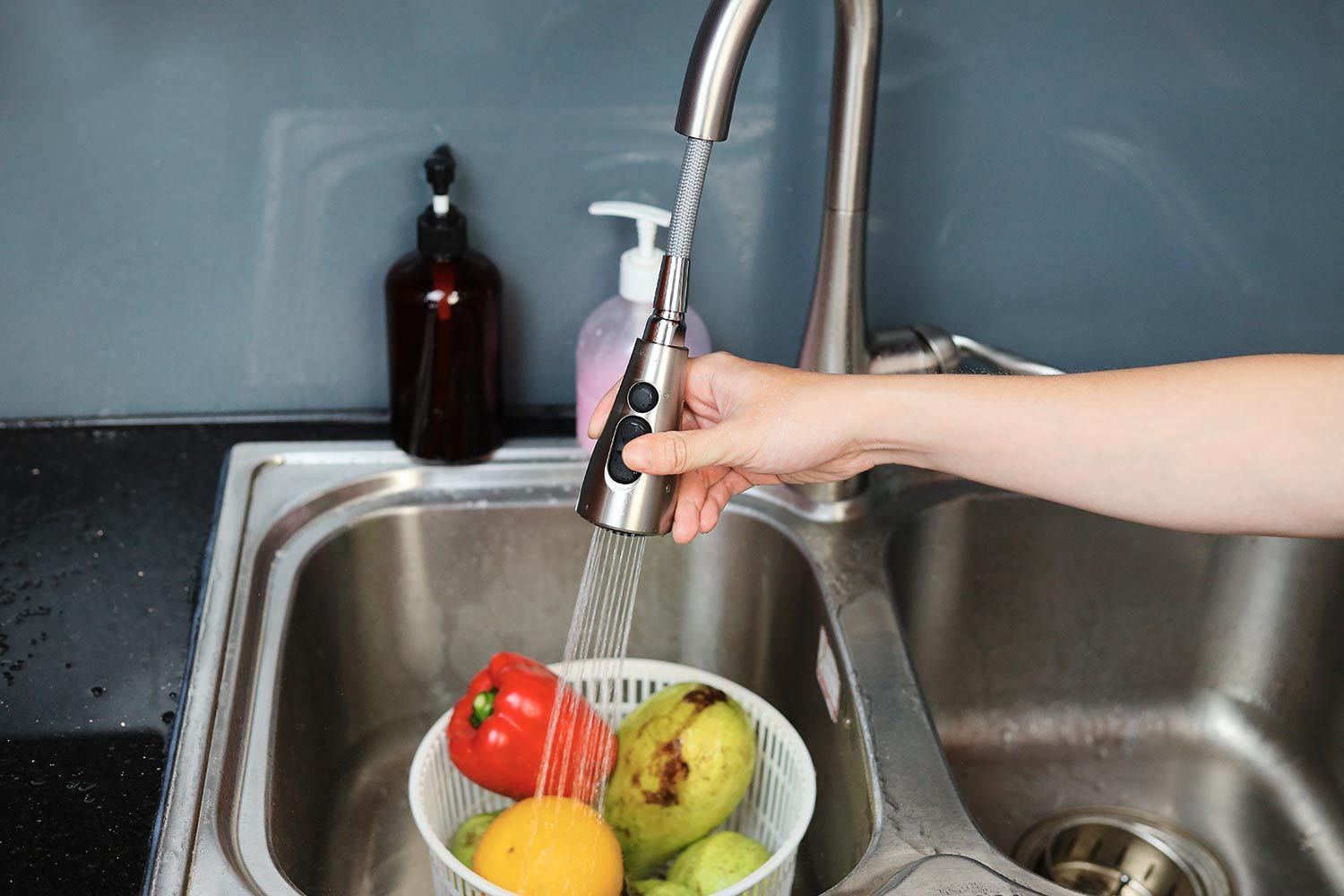 Hand pulls on the sprayer of a Moen Arbor 7594 kitchen faucet and washes fruits and vegetables in a kitchen sink.