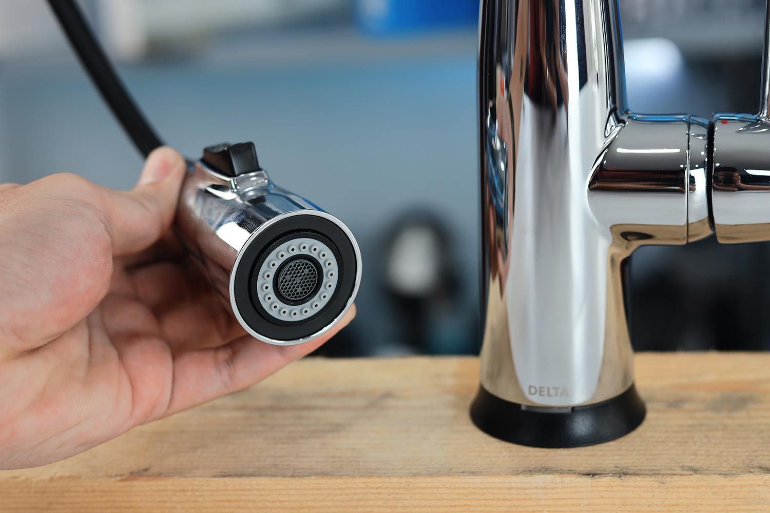 A hand holds a sprayer head of a Delta Faucet Essa 9113T-DST kitchen faucet installed on a wood platform.