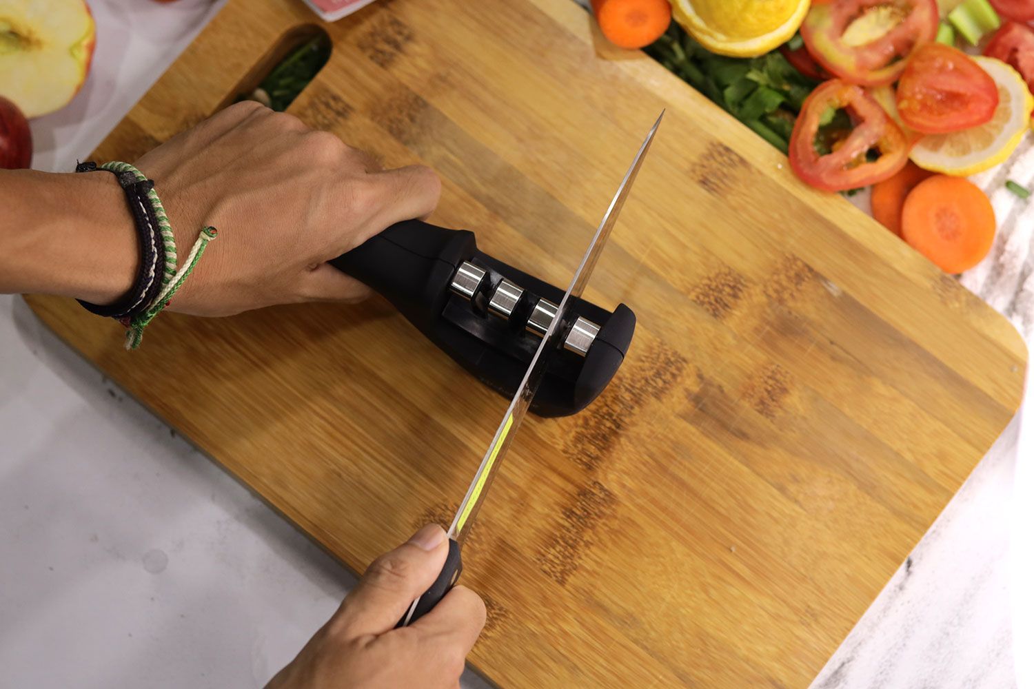 Top view of two hands sharpening a knife with the Kitchellence on cutting board, lemon and tomato slices in background