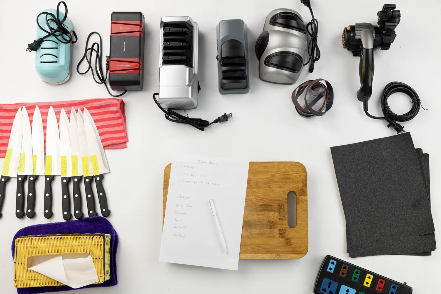 6 electric knife sharpeners, test knives on towel, paper and pen on cutting board, sandpapers, tissue box, extension socket.