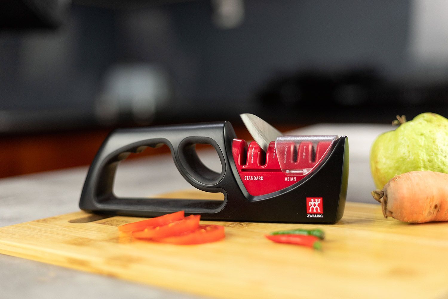 The Zwilling Henckels 4-Stage knife sharpener on a cutting board with chilli peppers, carrot, guava, tomato slices