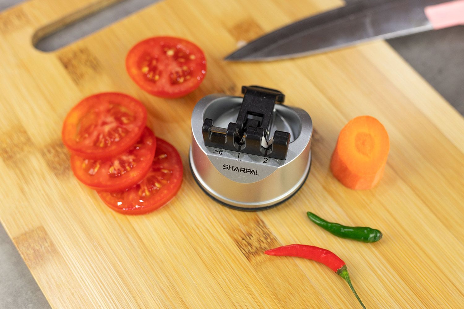 The Sharpal manual knife and scissor sharpener on cutting board, knife tip, chilli peppers, carrot slices, tomato slices