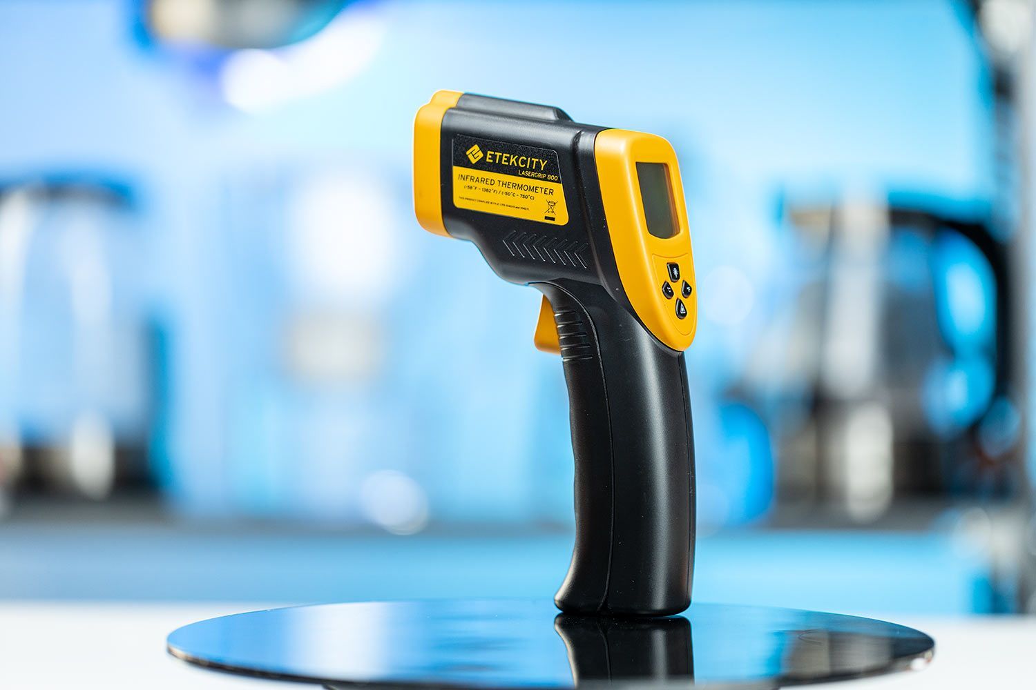 The Etekcity Lasergrip 800 Infrared Thermometer standing upright on a black turn table against a blurry, blue background.