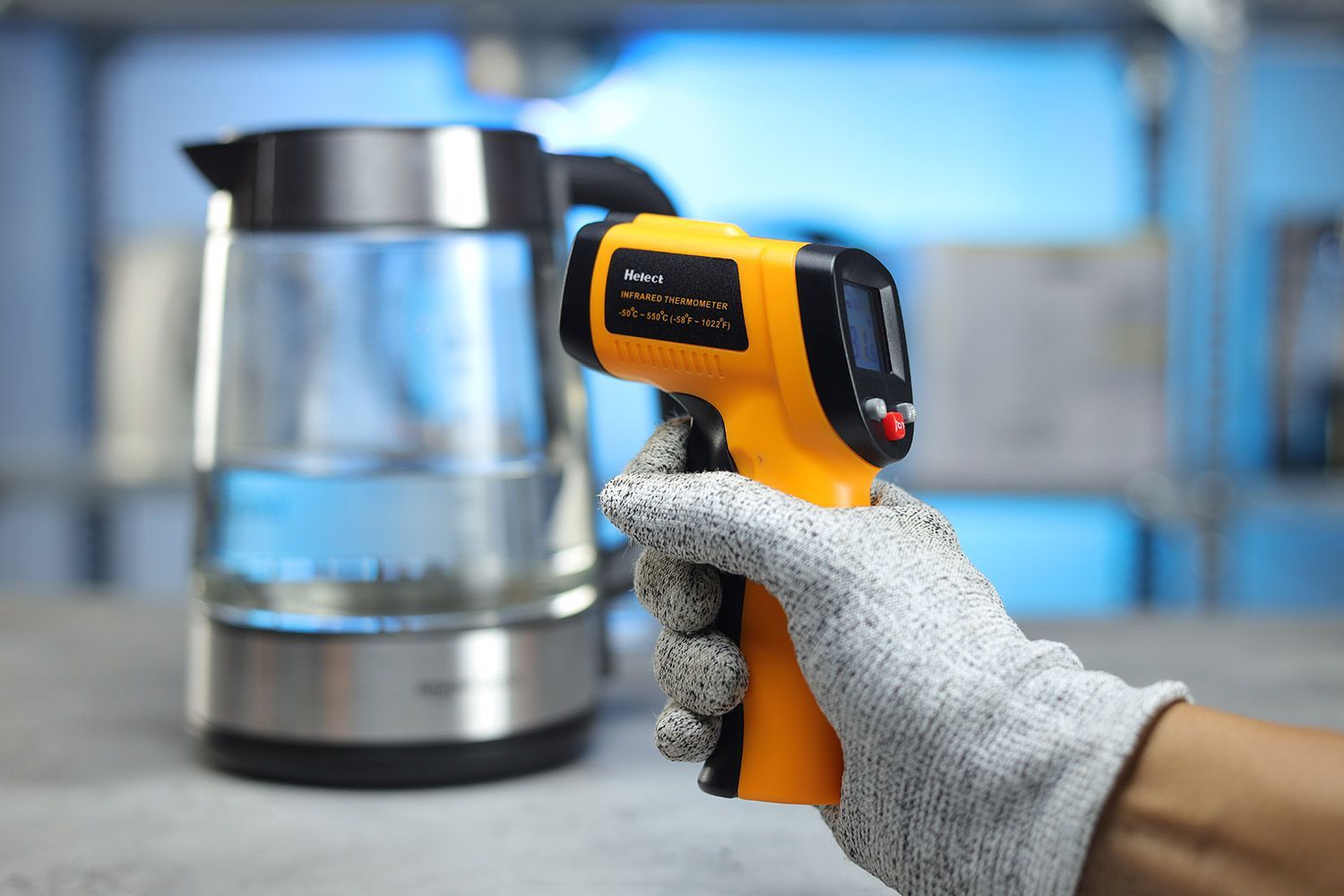 The Helect IR thermometer gun in the gloved hand of a reviewer against a blurry blue backdrop with a pitcher.