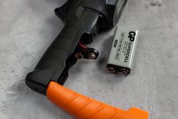 The 9V battery cell that comes with the Klein Tools IR1 laying on a concrete grey table. The thermometer itself is next to it. The battery compartment is opened.