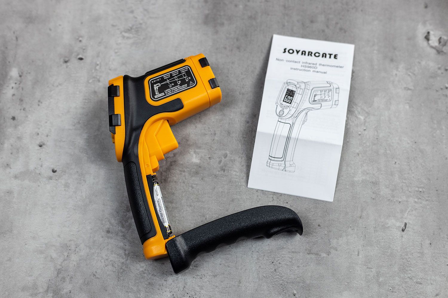 The battery compartment of the Sovarcate HS960D is opened. The user manual is to the right of the IR thermometer. Both are lying on a slate grey concrete table top.