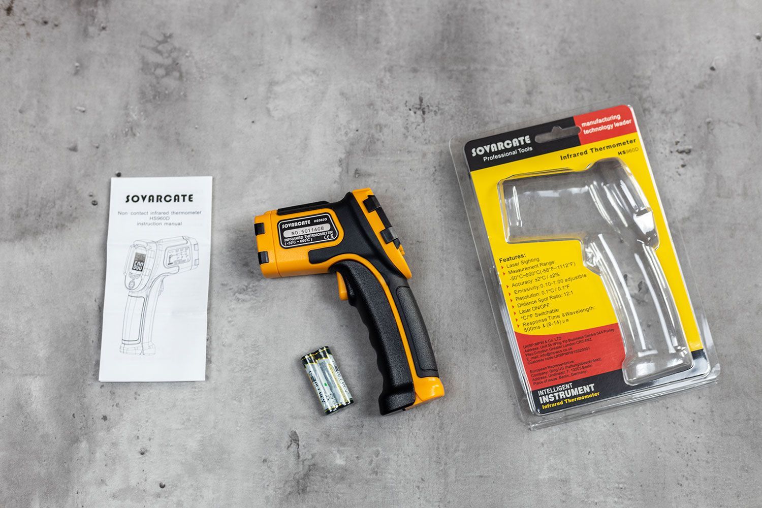 The Sovarcate HS960D in the center of the frame. To the left of the IR thermometer are the user manual and the batteries, and to the right is the plastic packaging.