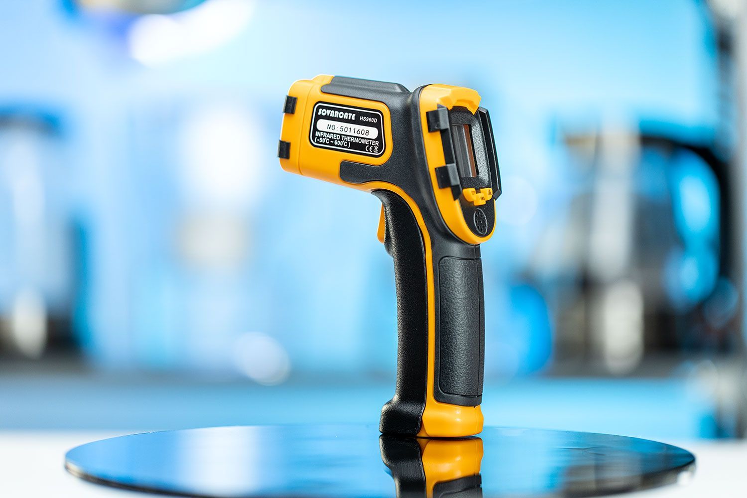 The SOVARCATE HS960D Infrared Thermometer standing upright on its handle on a turn table against a blurry blue backdrop.