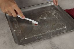 On a grey background, one hand brushing the silver baking pan of the Cuisinart TOA-60 Convection Toaster Oven Air Fryer with baking soda and water, the other holding a spray bottle of vinegar and water.