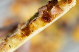 The cross-section of a toaster oven baked pizza slice showing the pizza crust. In the background is a 9-inch thick-crust meat pizza with cheese, onions, and green bell peppers on top inside a grooved silver baking tray.