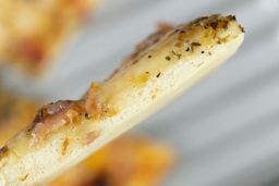 The cross-section of a toaster oven baked pizza slice showing the crust. In the background is a pizza inside a baking tray.