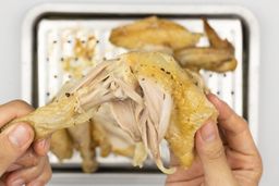 Two hands pulling apart a roasted chicken thigh. In the white background is the rest of the roasted chicken on a baking pan.