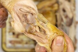 Two hands pulling apart a roasted chicken thigh. In the white background is the rest of the roasted chicken on a baking pan.